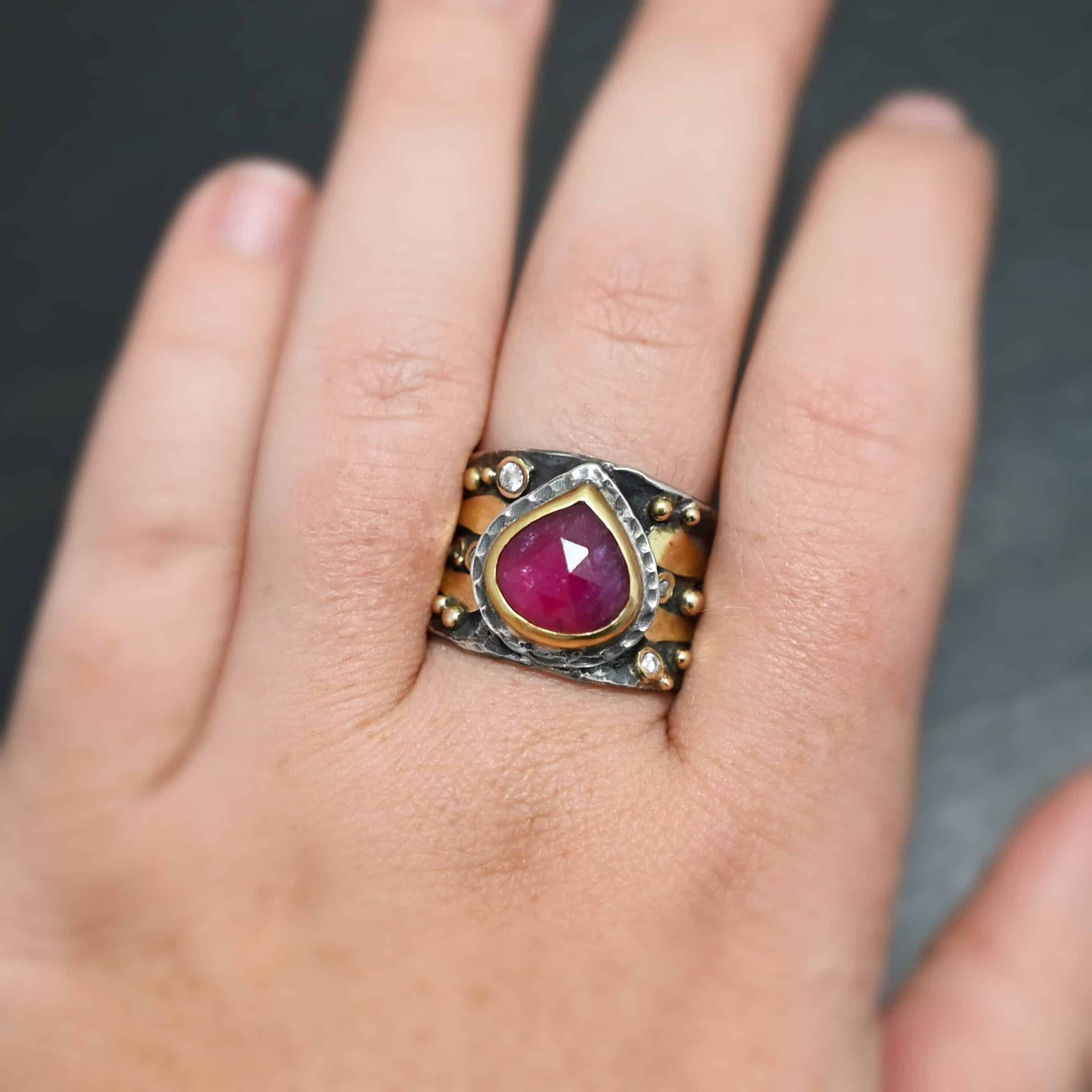 "Cherry Tree" Ring. Ring band is wide and sterling silver. There is a wide, pear shaped ruby in the center with hammered silver around it. There are two thicker bands of gold that wrap around the band. There are small diamonds and balls of gold scattered around the rest of the ring band. Ring is shown up close by a hand model.
