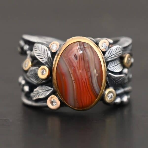 Close up of the "Season Of The Soul" Ring. This ring is made out of sterling silver, gold, teepee canyon agate, and moissanites. The center stone is the teepee canyon agate, which is primarily a mix of deep red colors. The agate is set in gold, as well as the moissanites that are scattered around the ring. There are silver leaves that are attached on both sides of the agate. The ring is made out of separate, but thick silver bands that connect to the agate setting to form the ring. There are balls of silver scattered around the ring as well. The ring is set against a dark background.
