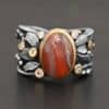 Close up of the "Season Of The Soul" Ring. This ring is made out of sterling silver, gold, teepee canyon agate, and moissanites. The center stone is the teepee canyon agate, which is primarily a mix of deep red colors. The agate is set in gold, as well as the moissanites that are scattered around the ring. There are silver leaves that are attached on both sides of the agate. The ring is made out of separate, but thick silver bands that connect to the agate setting to form the ring. There are balls of silver scattered around the ring as well. The ring is set against a dark background.