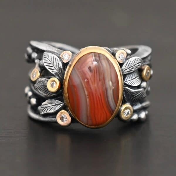 Close up of the "Season Of The Soul" Ring. This ring is made out of sterling silver, gold, teepee canyon agate, and moissanites. The center stone is the teepee canyon agate, which is primarily a mix of deep red colors. The agate is set in gold, as well as the moissanites that are scattered around the ring. There are silver leaves that are attached on both sides of the agate. The ring is made out of separate, but thick silver bands that connect to the agate setting to form the ring. There are balls of silver scattered around the ring as well. The ring is set against a dark background.