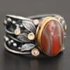 Close up of the "Season Of The Soul" Ring. This ring is made out of sterling silver, gold, teepee canyon agate, and moissanites. The center stone is the teepee canyon agate, which is primarily a mix of deep red colors. The agate is set in gold, as well as the moissanites that are scattered around the ring. There are silver leaves that are attached on both sides of the agate. The ring is made out of separate, but thick silver bands that connect to the agate setting to form the ring. There are balls of silver scattered around the ring as well. The ring is set against a dark background.