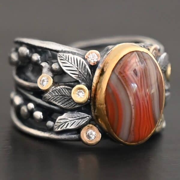 Close up of the "Season Of The Soul" Ring. This ring is made out of sterling silver, gold, teepee canyon agate, and moissanites. The center stone is the teepee canyon agate, which is primarily a mix of deep red colors. The agate is set in gold, as well as the moissanites that are scattered around the ring. There are silver leaves that are attached on both sides of the agate. The ring is made out of separate, but thick silver bands that connect to the agate setting to form the ring. There are balls of silver scattered around the ring as well. The ring is set against a dark background.