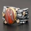 Close up of the "Season Of The Soul" Ring. This ring is made out of sterling silver, gold, teepee canyon agate, and moissanites. The center stone is the teepee canyon agate, which is primarily a mix of deep red colors. The agate is set in gold, as well as the moissanites that are scattered around the ring. There are silver leaves that are attached on both sides of the agate. The ring is made out of separate, but thick silver bands that connect to the agate setting to form the ring. There are balls of silver scattered around the ring as well. The ring is set against a dark background.