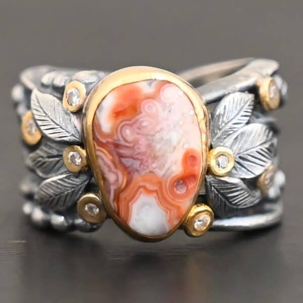 Close up of the "Maple" Ring. This ring is made out of sterling silver, gold, teepee canyon agate, and moissanites. The center stone is the teepee canyon agate, which is primarily a mix of orange, red, and white. The agate is set in gold, as well as the moissanites that are scattered around the ring. There are silver leaves that are attached on both sides of the agate. The ring is made out of separate, but thick silver bands that connect to the agate setting to form the ring. There are balls of silver scattered around the ring as well. The ring is set against a dark background.