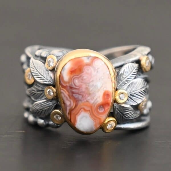Close up of the "Maple" Ring. This ring is made out of sterling silver, gold, teepee canyon agate, and moissanites. The center stone is the teepee canyon agate, which is primarily a mix of orange, red, and white. The agate is set in gold, as well as the moissanites that are scattered around the ring. There are silver leaves that are attached on both sides of the agate. The ring is made out of separate, but thick silver bands that connect to the agate setting to form the ring. There are balls of silver scattered around the ring as well. The ring is set against a dark background.