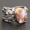 Close up of the "Maple" Ring. This ring is made out of sterling silver, gold, teepee canyon agate, and moissanites. The center stone is the teepee canyon agate, which is primarily a mix of orange, red, and white. The agate is set in gold, as well as the moissanites that are scattered around the ring. There are silver leaves that are attached on both sides of the agate. The ring is made out of separate, but thick silver bands that connect to the agate setting to form the ring. There are balls of silver scattered around the ring as well. The ring is set against a dark background.