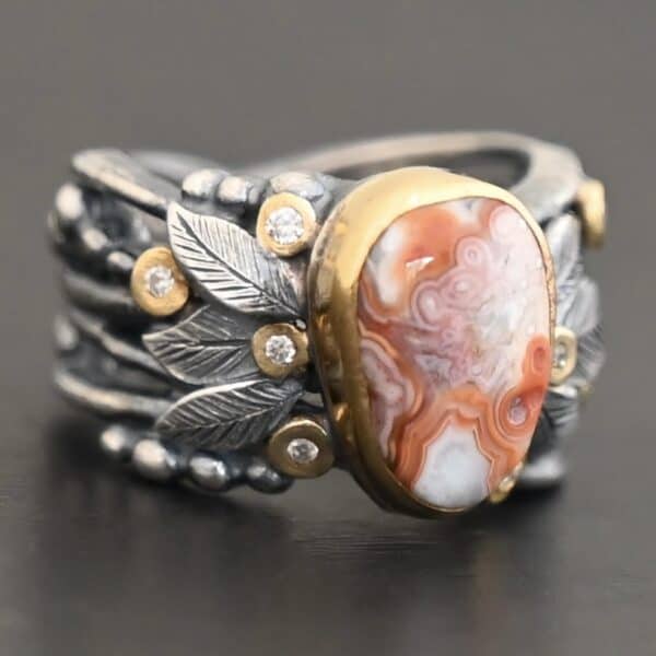 Close up of the "Maple" Ring. This ring is made out of sterling silver, gold, teepee canyon agate, and moissanites. The center stone is the teepee canyon agate, which is primarily a mix of orange, red, and white. The agate is set in gold, as well as the moissanites that are scattered around the ring. There are silver leaves that are attached on both sides of the agate. The ring is made out of separate, but thick silver bands that connect to the agate setting to form the ring. There are balls of silver scattered around the ring as well. The ring is set against a dark background.