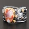 Close up of the "Maple" Ring. This ring is made out of sterling silver, gold, teepee canyon agate, and moissanites. The center stone is the teepee canyon agate, which is primarily a mix of orange, red, and white. The agate is set in gold, as well as the moissanites that are scattered around the ring. There are silver leaves that are attached on both sides of the agate. The ring is made out of separate, but thick silver bands that connect to the agate setting to form the ring. There are balls of silver scattered around the ring as well. The ring is set against a dark background.
