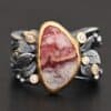 Close up of the "Rosemary" Ring. This ring is made out of sterling silver, gold, teepee canyon agate, and moissanites. The center stone is the teepee canyon agate, which is primarily a mix of maroon and white. The agate is set in gold, as well as the moissanites that are scattered around the ring. There are silver leaves that are attached on both sides of the agate. The ring is made out of separate, but thick silver bands that connect to the agate setting to form the ring. There are balls of silver scattered around the ring as well. The ring is set against a dark background.