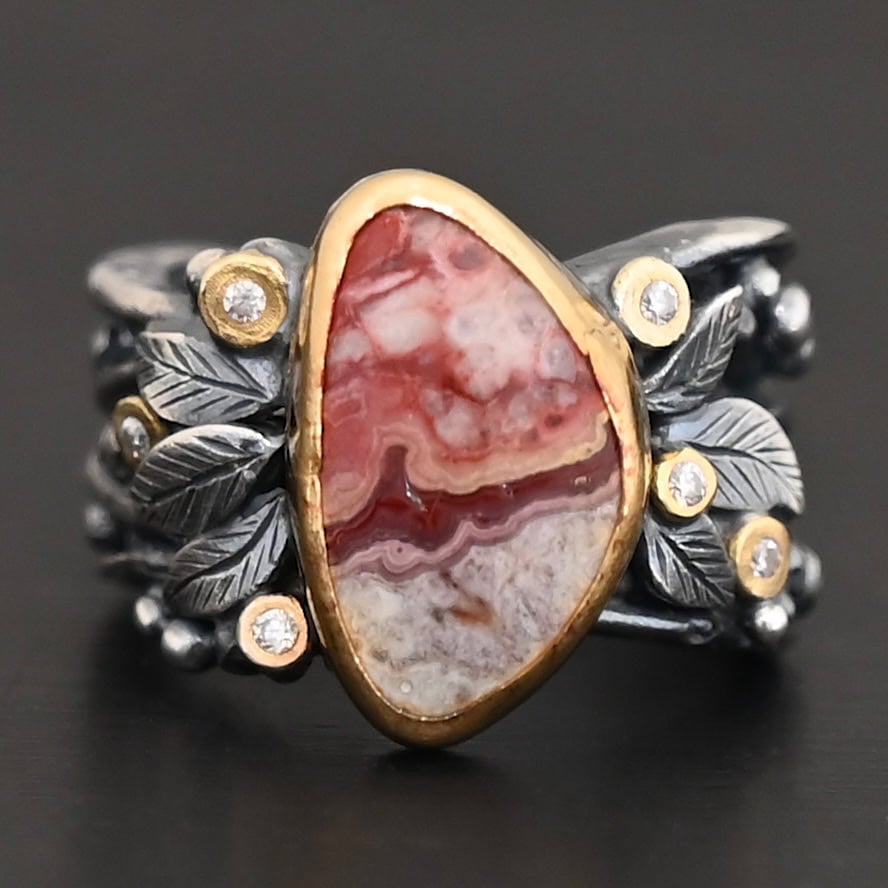 Close up of the "Rosemary" Ring. This ring is made out of sterling silver, gold, teepee canyon agate, and moissanites. The center stone is the teepee canyon agate, which is primarily a mix of maroon and white. The agate is set in gold, as well as the moissanites that are scattered around the ring. There are silver leaves that are attached on both sides of the agate. The ring is made out of separate, but thick silver bands that connect to the agate setting to form the ring. There are balls of silver scattered around the ring as well. The ring is set against a dark background.