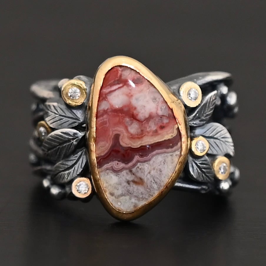 Close up of the "Rosemary" Ring. This ring is made out of sterling silver, gold, teepee canyon agate, and moissanites. The center stone is the teepee canyon agate, which is primarily a mix of maroon and white. The agate is set in gold, as well as the moissanites that are scattered around the ring. There are silver leaves that are attached on both sides of the agate. The ring is made out of separate, but thick silver bands that connect to the agate setting to form the ring. There are balls of silver scattered around the ring as well. The ring is set against a dark background.