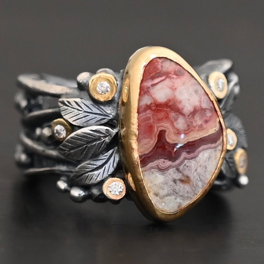 Close up of the "Rosemary" Ring. This ring is made out of sterling silver, gold, teepee canyon agate, and moissanites. The center stone is the teepee canyon agate, which is primarily a mix of maroon and white. The agate is set in gold, as well as the moissanites that are scattered around the ring. There are silver leaves that are attached on both sides of the agate. The ring is made out of separate, but thick silver bands that connect to the agate setting to form the ring. There are balls of silver scattered around the ring as well. The ring is set against a dark background.