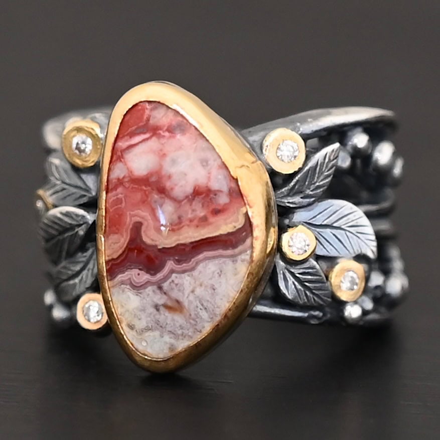 Close up of the "Rosemary" Ring. This ring is made out of sterling silver, gold, teepee canyon agate, and moissanites. The center stone is the teepee canyon agate, which is primarily a mix of maroon and white. The agate is set in gold, as well as the moissanites that are scattered around the ring. There are silver leaves that are attached on both sides of the agate. The ring is made out of separate, but thick silver bands that connect to the agate setting to form the ring. There are balls of silver scattered around the ring as well. The ring is set against a dark background.