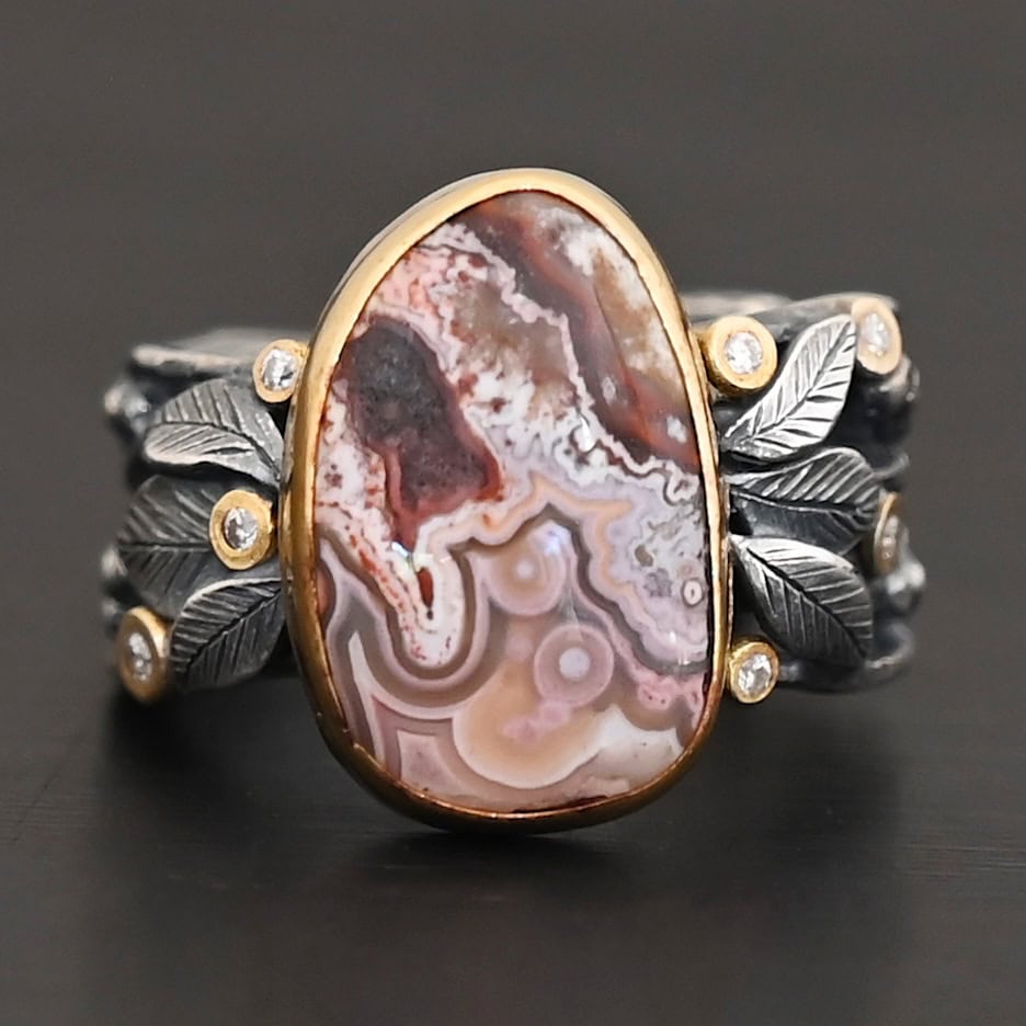 Close up of the "Cinnamon" Ring. This ring is made out of sterling silver, gold, teepee canyon agate, and moissanites. The center stone is the teepee canyon agate, which is primarily a mix of maroon and white. The agate is set in gold, as well as the moissanites that are scattered around the ring. There are silver leaves that are attached on both sides of the agate. The ring is made out of separate, but thick silver bands that connect to the agate setting to form the ring. There are balls of silver scattered around the ring as well. The ring is set against a dark background.
