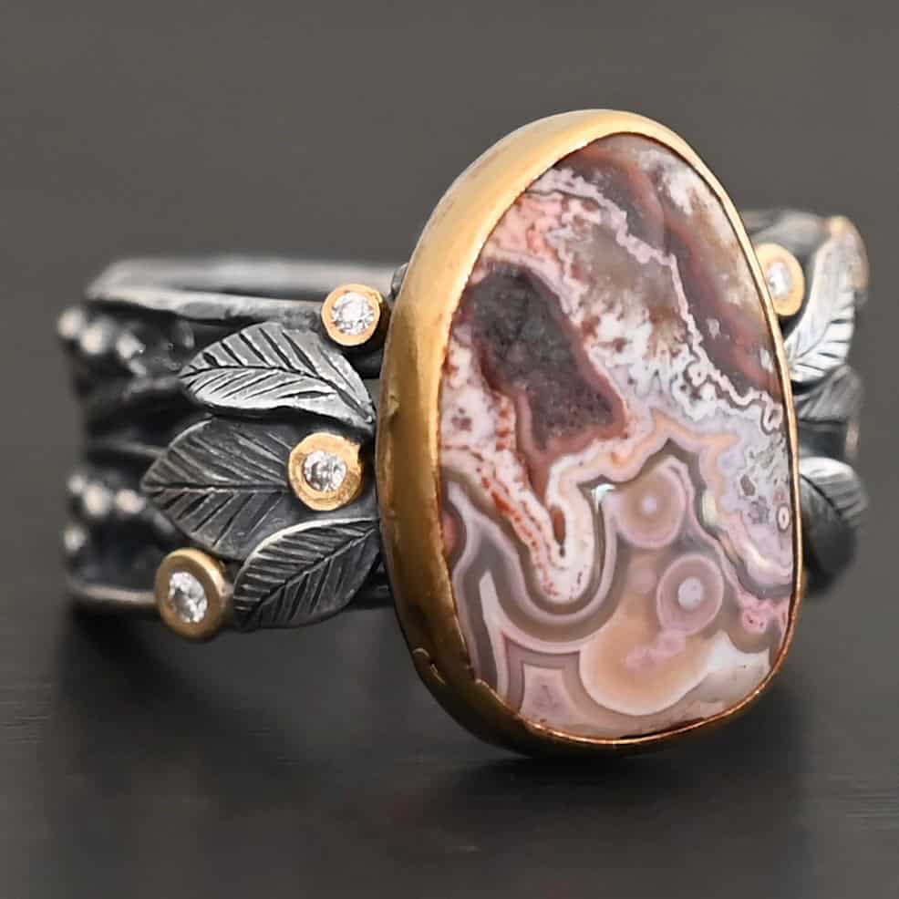 Close up of the "Cinnamon" Ring. This ring is made out of sterling silver, gold, teepee canyon agate, and moissanites. The center stone is the teepee canyon agate, which is primarily a mix of maroon and white. The agate is set in gold, as well as the moissanites that are scattered around the ring. There are silver leaves that are attached on both sides of the agate. The ring is made out of separate, but thick silver bands that connect to the agate setting to form the ring. There are balls of silver scattered around the ring as well. The ring is set against a dark background.