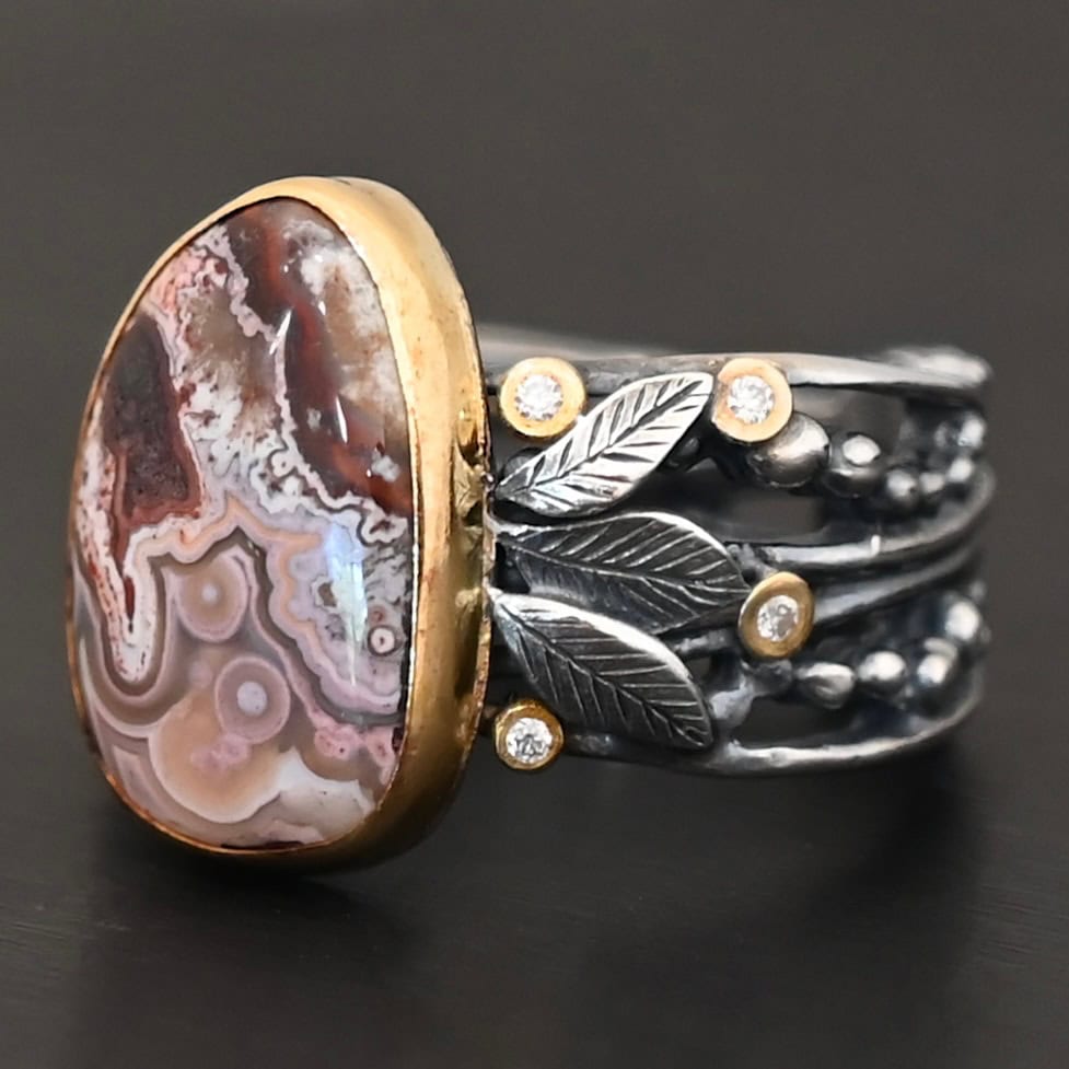 Close up of the "Cinnamon" Ring. This ring is made out of sterling silver, gold, teepee canyon agate, and moissanites. The center stone is the teepee canyon agate, which is primarily a mix of maroon and white. The agate is set in gold, as well as the moissanites that are scattered around the ring. There are silver leaves that are attached on both sides of the agate. The ring is made out of separate, but thick silver bands that connect to the agate setting to form the ring. There are balls of silver scattered around the ring as well. The ring is set against a dark background.