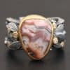 Close up of the "Saige" Ring. This ring is made out of sterling silver, gold, teepee canyon agate, and moissanites. The center stone is the teepee canyon agate, which is primarily a mix of maroon and white. The agate is set in gold, as well as the moissanites that are scattered around the ring. There are silver leaves that are attached on both sides of the agate. The ring is made out of separate, but thick silver bands that connect to the agate setting to form the ring. There are balls of silver scattered around the ring as well. The ring is set against a dark background.