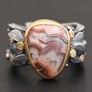 Close up of the "Saige" Ring. This ring is made out of sterling silver, gold, teepee canyon agate, and moissanites. The center stone is the teepee canyon agate, which is primarily a mix of maroon and white. The agate is set in gold, as well as the moissanites that are scattered around the ring. There are silver leaves that are attached on both sides of the agate. The ring is made out of separate, but thick silver bands that connect to the agate setting to form the ring. There are balls of silver scattered around the ring as well. The ring is set against a dark background.