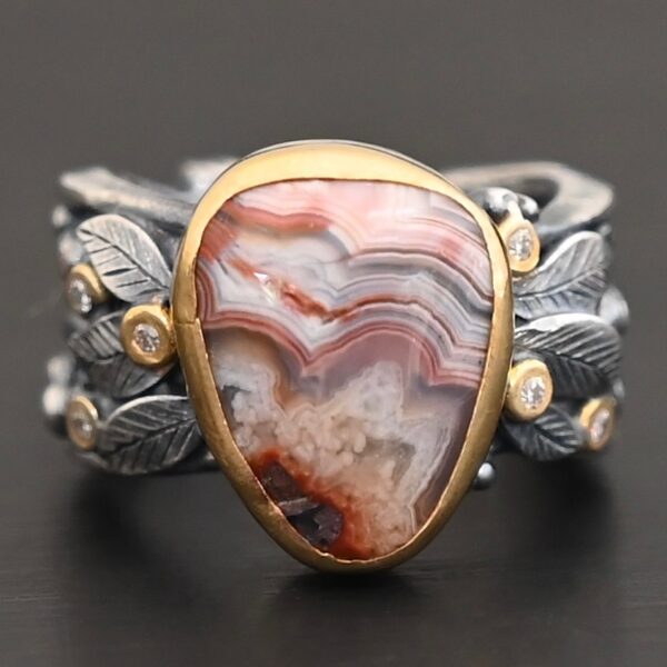 Close up of the "Saige" Ring. This ring is made out of sterling silver, gold, teepee canyon agate, and moissanites. The center stone is the teepee canyon agate, which is primarily a mix of maroon and white. The agate is set in gold, as well as the moissanites that are scattered around the ring. There are silver leaves that are attached on both sides of the agate. The ring is made out of separate, but thick silver bands that connect to the agate setting to form the ring. There are balls of silver scattered around the ring as well. The ring is set against a dark background.