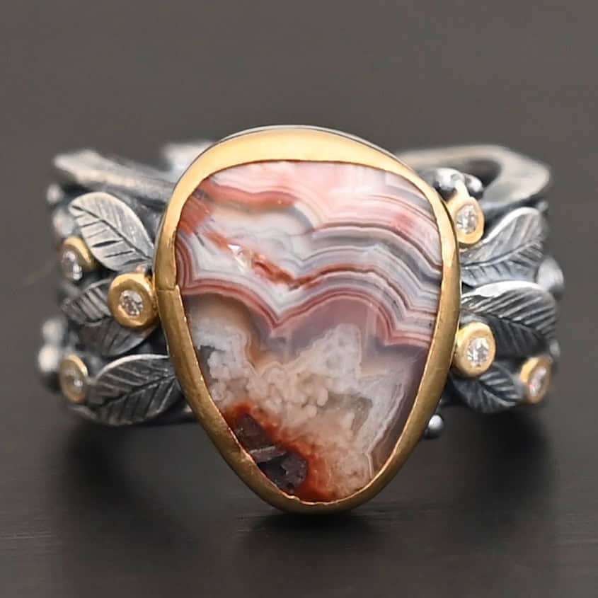 Close up of the "Saige" Ring. This ring is made out of sterling silver, gold, teepee canyon agate, and moissanites. The center stone is the teepee canyon agate, which is primarily a mix of maroon and white. The agate is set in gold, as well as the moissanites that are scattered around the ring. There are silver leaves that are attached on both sides of the agate. The ring is made out of separate, but thick silver bands that connect to the agate setting to form the ring. There are balls of silver scattered around the ring as well. The ring is set against a dark background.