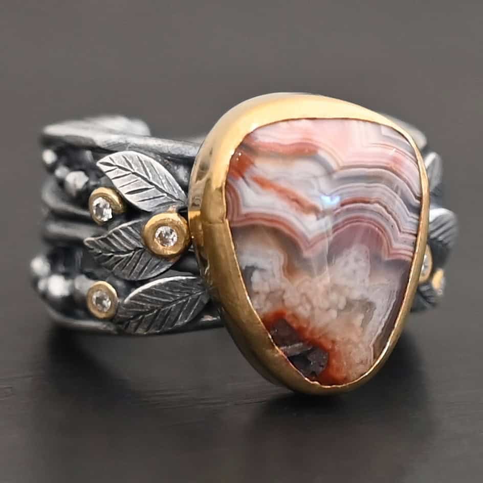 Close up of the "Saige" Ring. This ring is made out of sterling silver, gold, teepee canyon agate, and moissanites. The center stone is the teepee canyon agate, which is primarily a mix of maroon and white. The agate is set in gold, as well as the moissanites that are scattered around the ring. There are silver leaves that are attached on both sides of the agate. The ring is made out of separate, but thick silver bands that connect to the agate setting to form the ring. There are balls of silver scattered around the ring as well. The ring is set against a dark background.