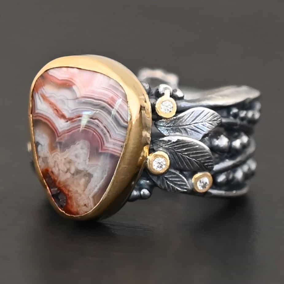 Close up of the "Saige" Ring. This ring is made out of sterling silver, gold, teepee canyon agate, and moissanites. The center stone is the teepee canyon agate, which is primarily a mix of maroon and white. The agate is set in gold, as well as the moissanites that are scattered around the ring. There are silver leaves that are attached on both sides of the agate. The ring is made out of separate, but thick silver bands that connect to the agate setting to form the ring. There are balls of silver scattered around the ring as well. The ring is set against a dark background.