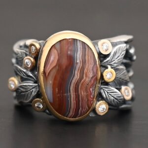 Close up of the "Mellow Season" Ring. This ring is made out of sterling silver, gold, teepee canyon agate, and moissanites. The center stone is the teepee canyon agate, which is primarily a mix of deep red and white lines. The agate is set in gold, as well as the moissanites that are scattered around the ring. There are silver leaves that are attached on both sides of the agate. The ring is made out of separate, but thick silver bands that connect to the agate setting to form the ring. There are balls of silver scattered around the ring as well. The ring is set against a dark background.