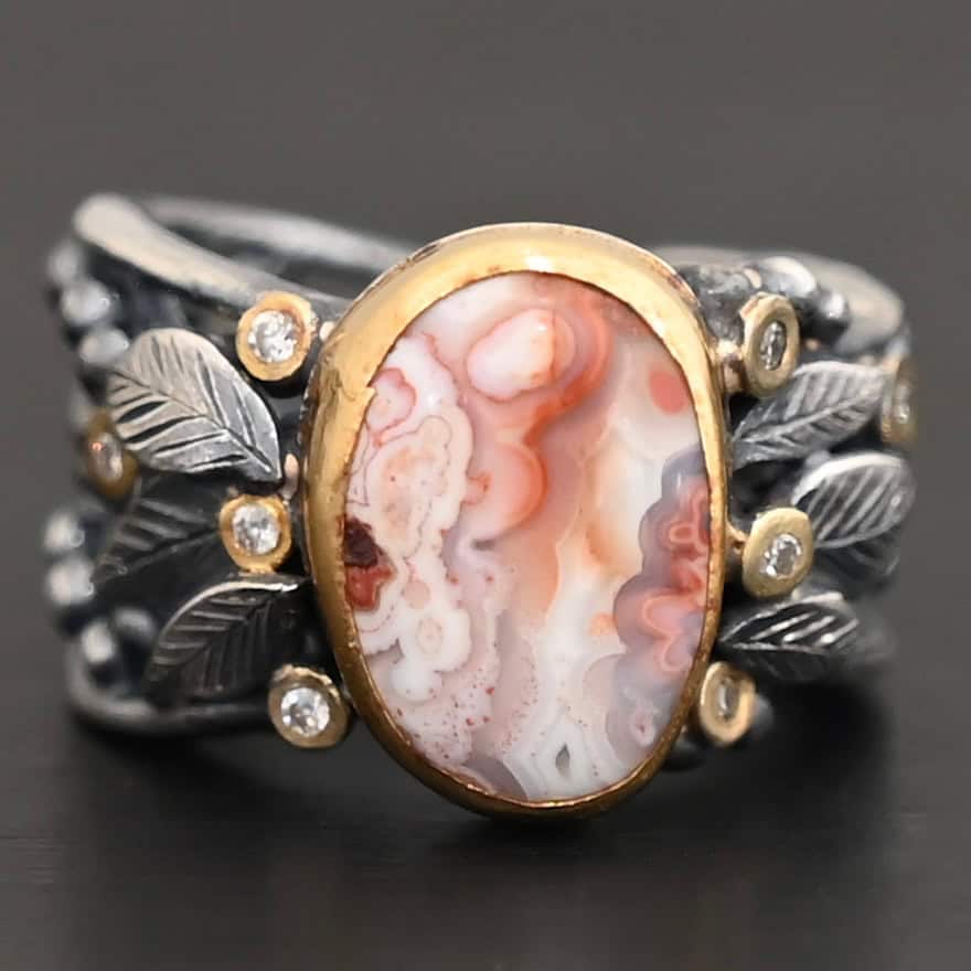 Close up of the "Turn The Page" Ring. This ring is made out of sterling silver, gold, teepee canyon agate, and moissanites. The center stone is the teepee canyon agate, which is primarily a mix of orange, red, and white. The agate is set in gold, as well as the moissanites that are scattered around the ring. There are silver leaves that are attached on both sides of the agate. The ring is made out of separate, but thick silver bands that connect to the agate setting to form the ring. There are balls of silver scattered around the ring as well. The ring is set against a dark background.