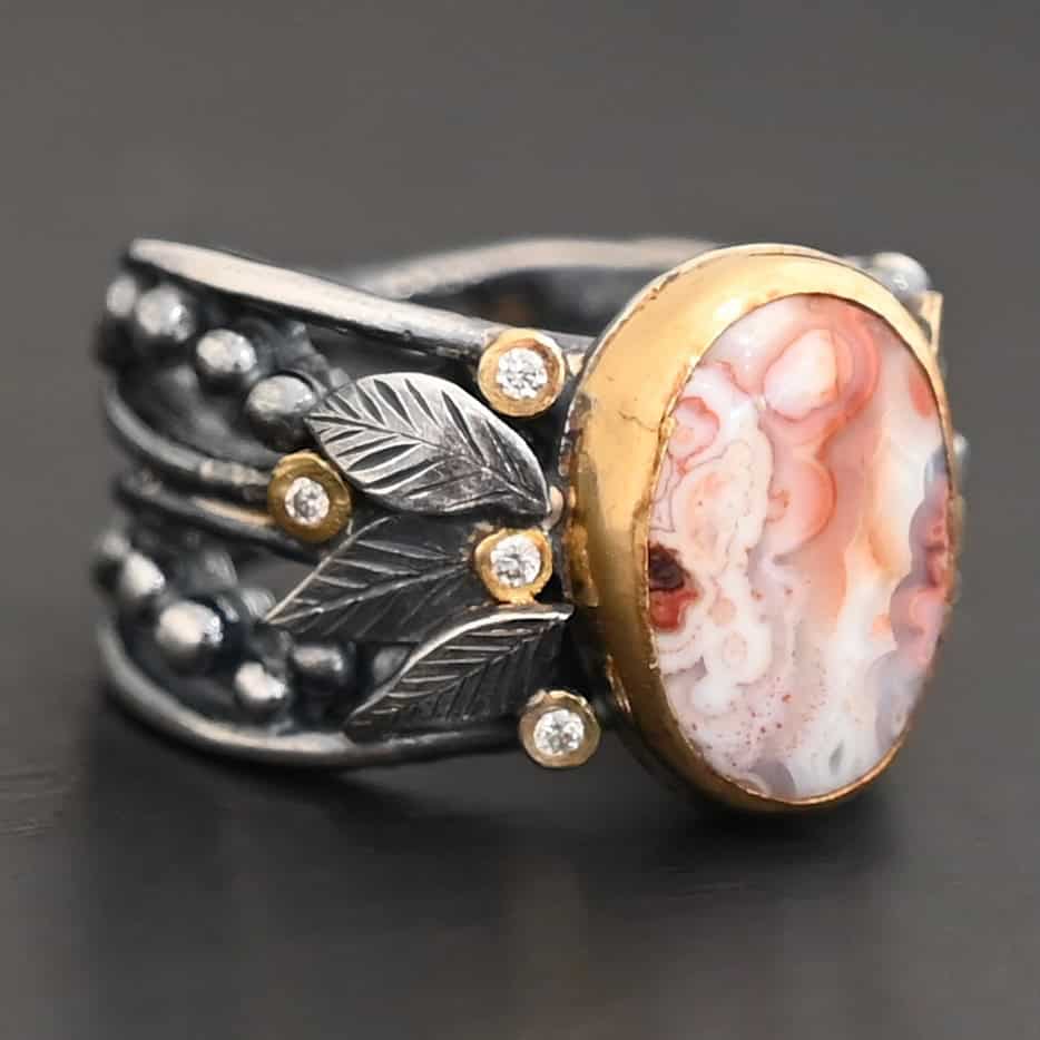 Close up of the "Turn The Page" Ring. This ring is made out of sterling silver, gold, teepee canyon agate, and moissanites. The center stone is the teepee canyon agate, which is primarily a mix of orange, red, and white. The agate is set in gold, as well as the moissanites that are scattered around the ring. There are silver leaves that are attached on both sides of the agate. The ring is made out of separate, but thick silver bands that connect to the agate setting to form the ring. There are balls of silver scattered around the ring as well. The ring is set against a dark background.