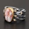 Close up of the "Turn The Page" Ring. This ring is made out of sterling silver, gold, teepee canyon agate, and moissanites. The center stone is the teepee canyon agate, which is primarily a mix of orange, red, and white. The agate is set in gold, as well as the moissanites that are scattered around the ring. There are silver leaves that are attached on both sides of the agate. The ring is made out of separate, but thick silver bands that connect to the agate setting to form the ring. There are balls of silver scattered around the ring as well. The ring is set against a dark background.