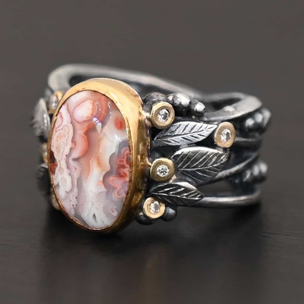 Close up of the "Turn The Page" Ring. This ring is made out of sterling silver, gold, teepee canyon agate, and moissanites. The center stone is the teepee canyon agate, which is primarily a mix of orange, red, and white. The agate is set in gold, as well as the moissanites that are scattered around the ring. There are silver leaves that are attached on both sides of the agate. The ring is made out of separate, but thick silver bands that connect to the agate setting to form the ring. There are balls of silver scattered around the ring as well. The ring is set against a dark background.