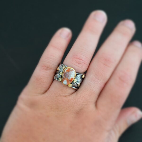 Close up of the "Maple" Ring. This ring is made out of sterling silver, gold, teepee canyon agate, and moissanites. The center stone is the teepee canyon agate, which is primarily a mix of orange, red, and white. The agate is set in gold, as well as the moissanites that are scattered around the ring. There are silver leaves that are attached on both sides of the agate. The ring is made out of separate, but thick silver bands that connect to the agate setting to form the ring. There are balls of silver scattered around the ring as well. Ring is worn up close by a hand model.