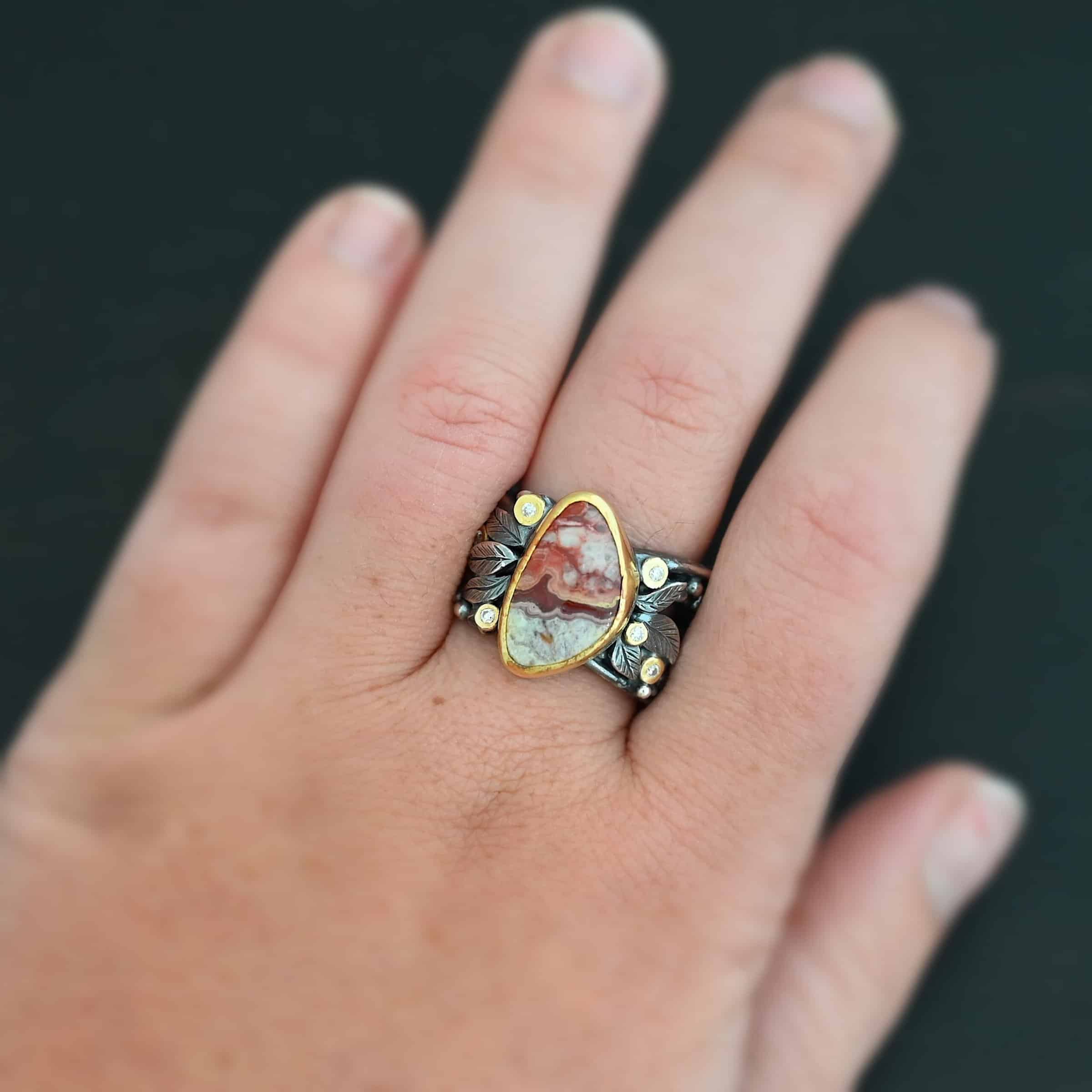 Close up of the "Rosemary" Ring. This ring is made out of sterling silver, gold, teepee canyon agate, and moissanites. The center stone is the teepee canyon agate, which is primarily a mix of maroon and white. The agate is set in gold, as well as the moissanites that are scattered around the ring. There are silver leaves that are attached on both sides of the agate. The ring is made out of separate, but thick silver bands that connect to the agate setting to form the ring. There are balls of silver scattered around the ring as well. Ring is worn up close by a hand model.