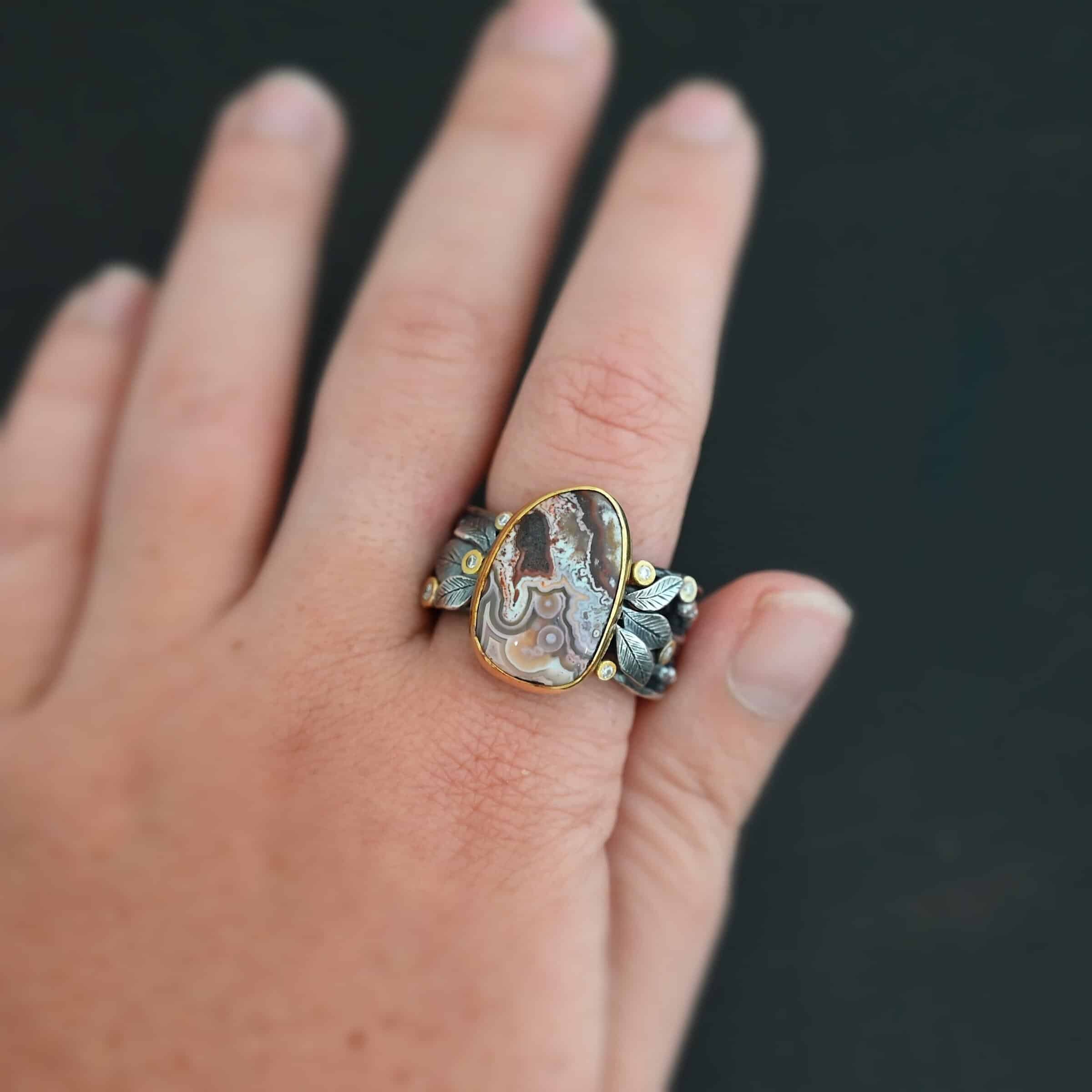 Close up of the "Cinnamon" Ring. This ring is made out of sterling silver, gold, teepee canyon agate, and moissanites. The center stone is the teepee canyon agate, which is primarily a mix of maroon and white. The agate is set in gold, as well as the moissanites that are scattered around the ring. There are silver leaves that are attached on both sides of the agate. The ring is made out of separate, but thick silver bands that connect to the agate setting to form the ring. There are balls of silver scattered around the ring as well. Ring is worn up close by a hand model.
