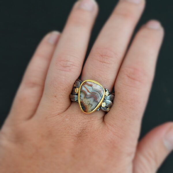 Close up of the "Saige" Ring. This ring is made out of sterling silver, gold, teepee canyon agate, and moissanites. The center stone is the teepee canyon agate, which is primarily a mix of maroon and white. The agate is set in gold, as well as the moissanites that are scattered around the ring. There are silver leaves that are attached on both sides of the agate. The ring is made out of separate, but thick silver bands that connect to the agate setting to form the ring. There are balls of silver scattered around the ring as well. Ring is worn up close by a hand model.