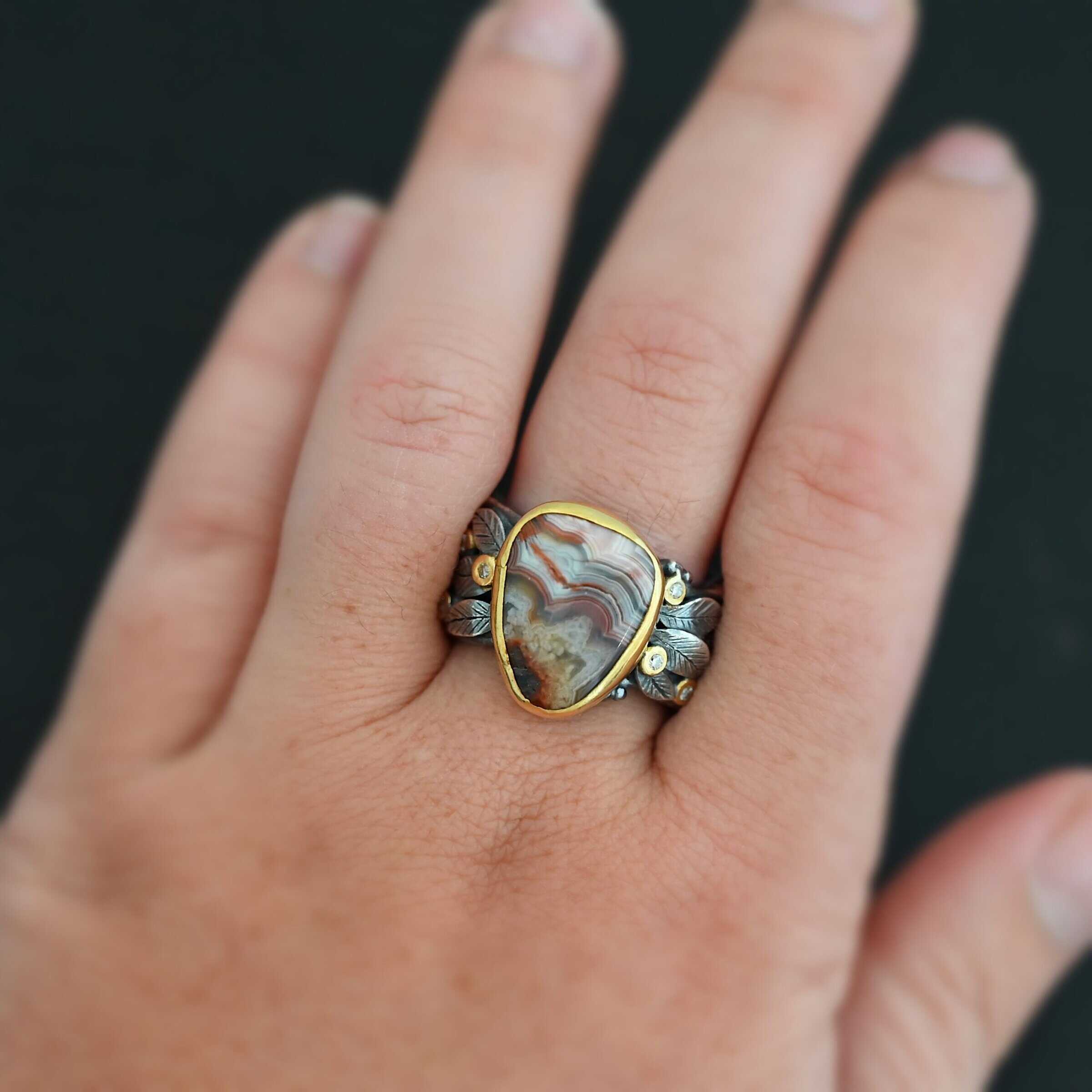 Close up of the "Saige" Ring. This ring is made out of sterling silver, gold, teepee canyon agate, and moissanites. The center stone is the teepee canyon agate, which is primarily a mix of maroon and white. The agate is set in gold, as well as the moissanites that are scattered around the ring. There are silver leaves that are attached on both sides of the agate. The ring is made out of separate, but thick silver bands that connect to the agate setting to form the ring. There are balls of silver scattered around the ring as well. Ring is worn up close by a hand model.