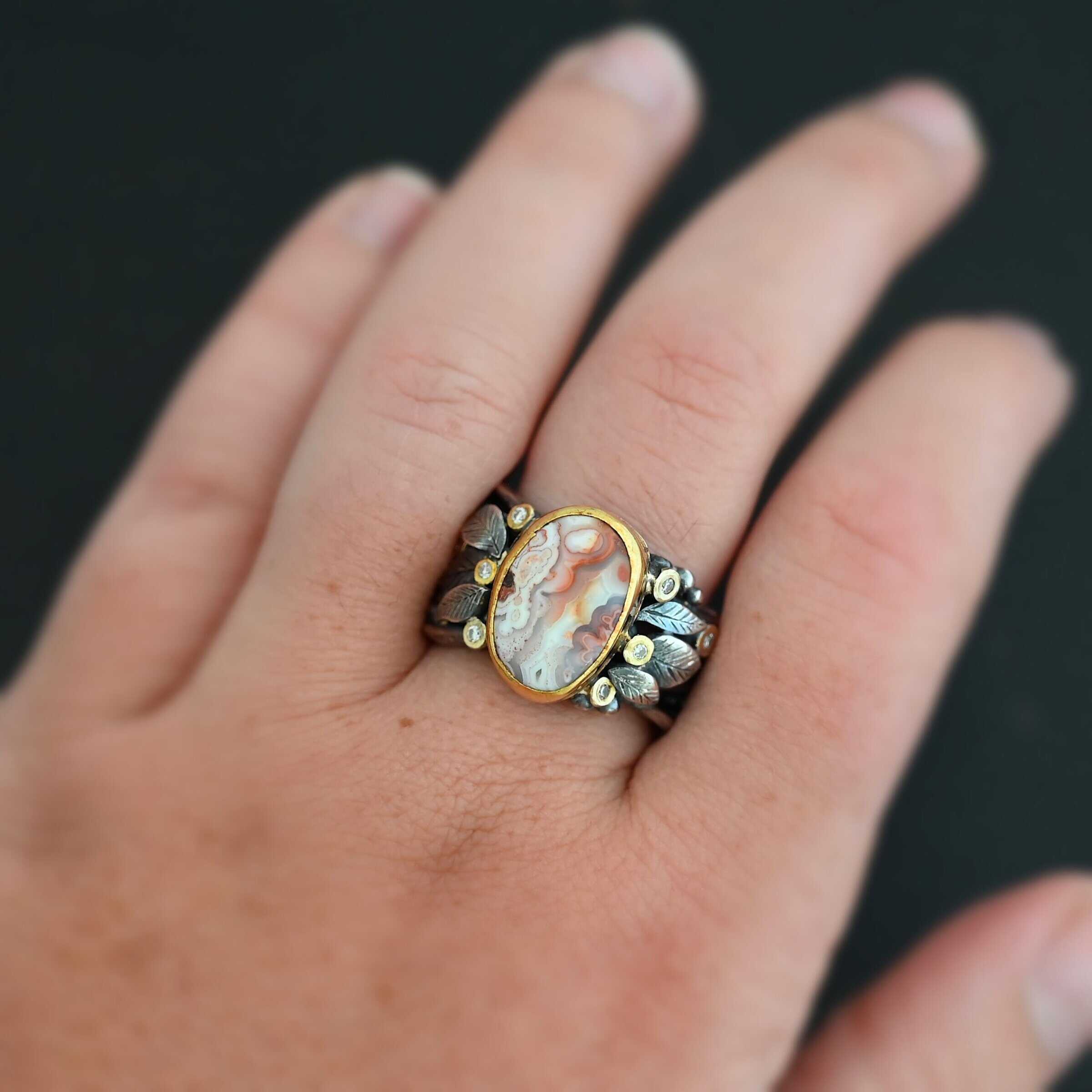 Close up of the "Turn The Page" Ring. This ring is made out of sterling silver, gold, teepee canyon agate, and moissanites. The center stone is the teepee canyon agate, which is primarily a mix of orange, red, and white. The agate is set in gold, as well as the moissanites that are scattered around the ring. There are silver leaves that are attached on both sides of the agate. The ring is made out of separate, but thick silver bands that connect to the agate setting to form the ring. There are balls of silver scattered around the ring as well. Ring is worn up close by a hand model.