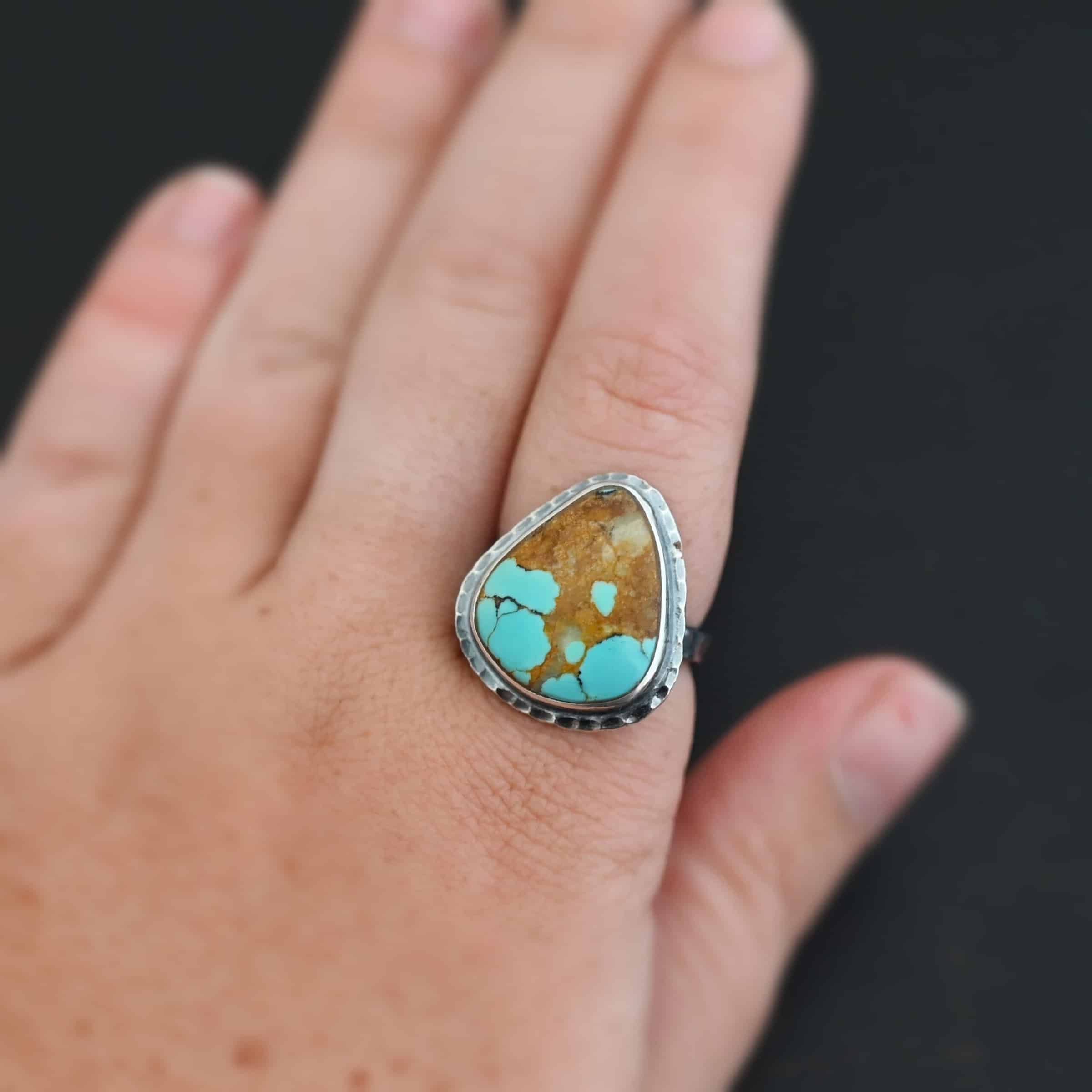 "Arleen" Ring. There is a mostly oval turquoise in the center with hammered sterling silver around it. Ring is shown up close on a model's hand.
