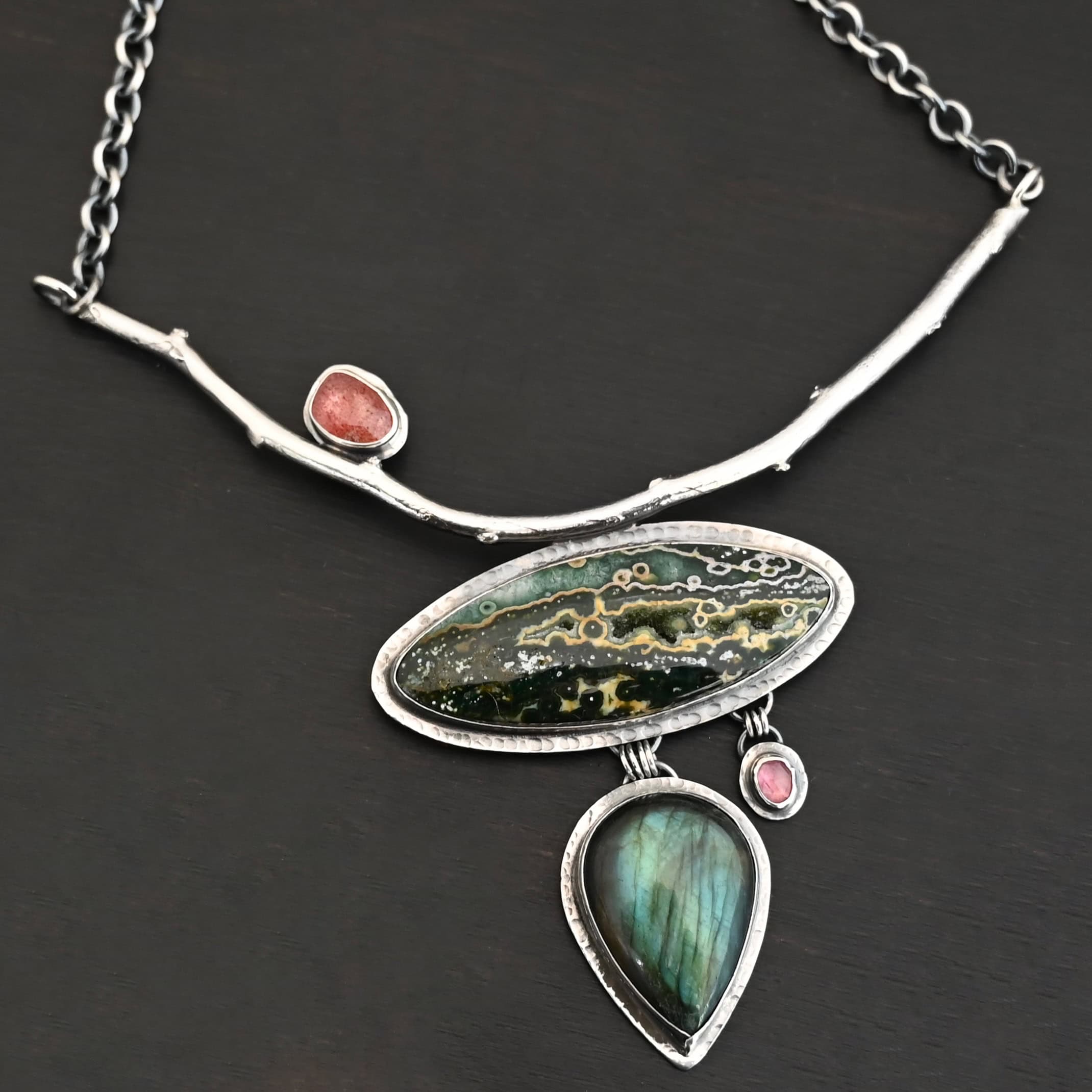 In The Darkness, There Is Light Necklace "In The Darkness, There is Light" Necklace. There is a large, curved, sterling silver casted twig. Soldered beneath it is a horizontal, oval ocean jasper. There is a teardrop labradorite hanging beneath that, as well as a small pink tourmaline next to that. There is another oval stone soldered to the top left side of the branch.