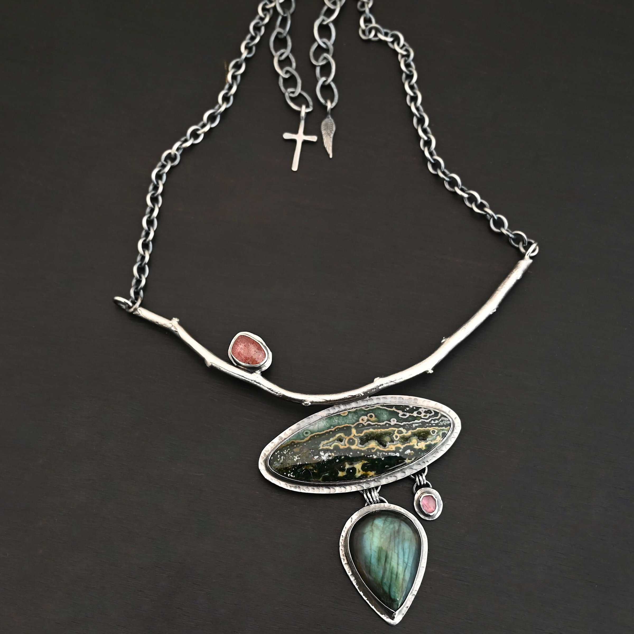 In The Darkness, There Is Light Necklace "In The Darkness, There is Light" Necklace. There is a large, curved, sterling silver casted twig. Soldered beneath it is a horizontal, oval ocean jasper. There is a teardrop labradorite hanging beneath that, as well as a small pink tourmaline next to that. There is another oval stone soldered to the top left side of the branch.