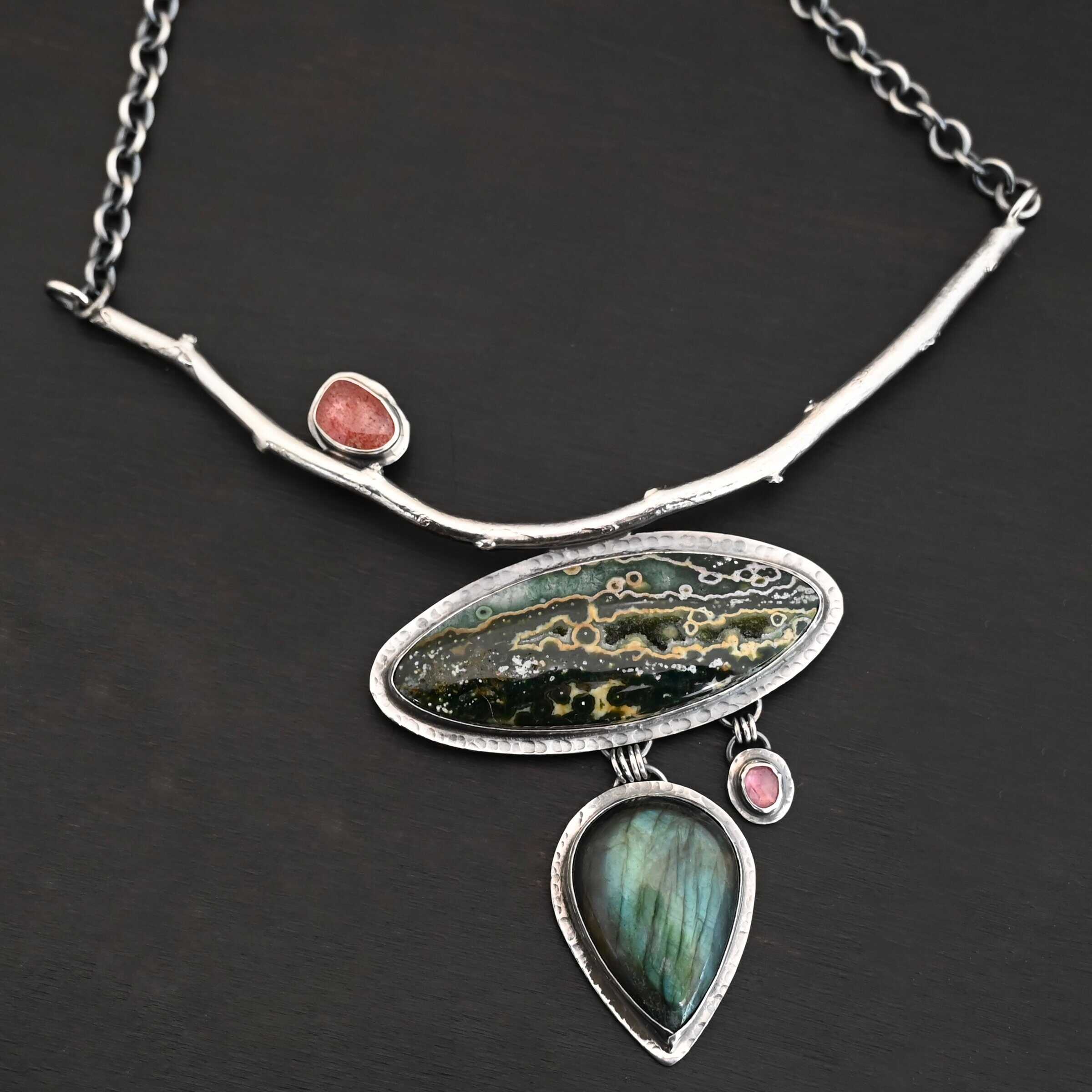 In The Darkness, There Is Light Necklace "In The Darkness, There is Light" Necklace. There is a large, curved, sterling silver casted twig. Soldered beneath it is a horizontal, oval ocean jasper. There is a teardrop labradorite hanging beneath that, as well as a small pink tourmaline next to that. There is another oval stone soldered to the top left side of the branch.