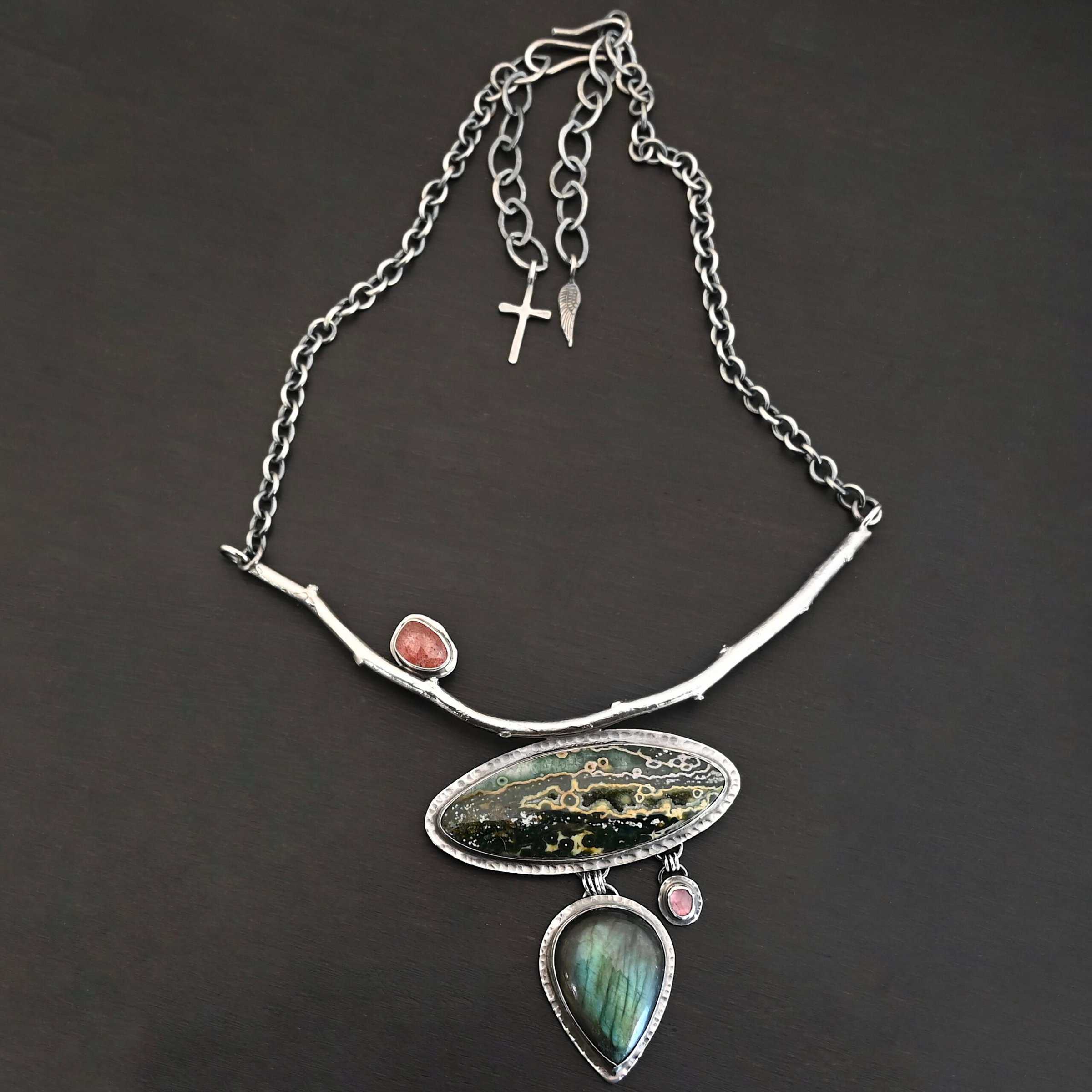 In The Darkness, There Is Light Necklace "In The Darkness, There is Light" Necklace. There is a large, curved, sterling silver casted twig. Soldered beneath it is a horizontal, oval ocean jasper. There is a teardrop labradorite hanging beneath that, as well as a small pink tourmaline next to that. There is another oval stone soldered to the top left side of the branch.