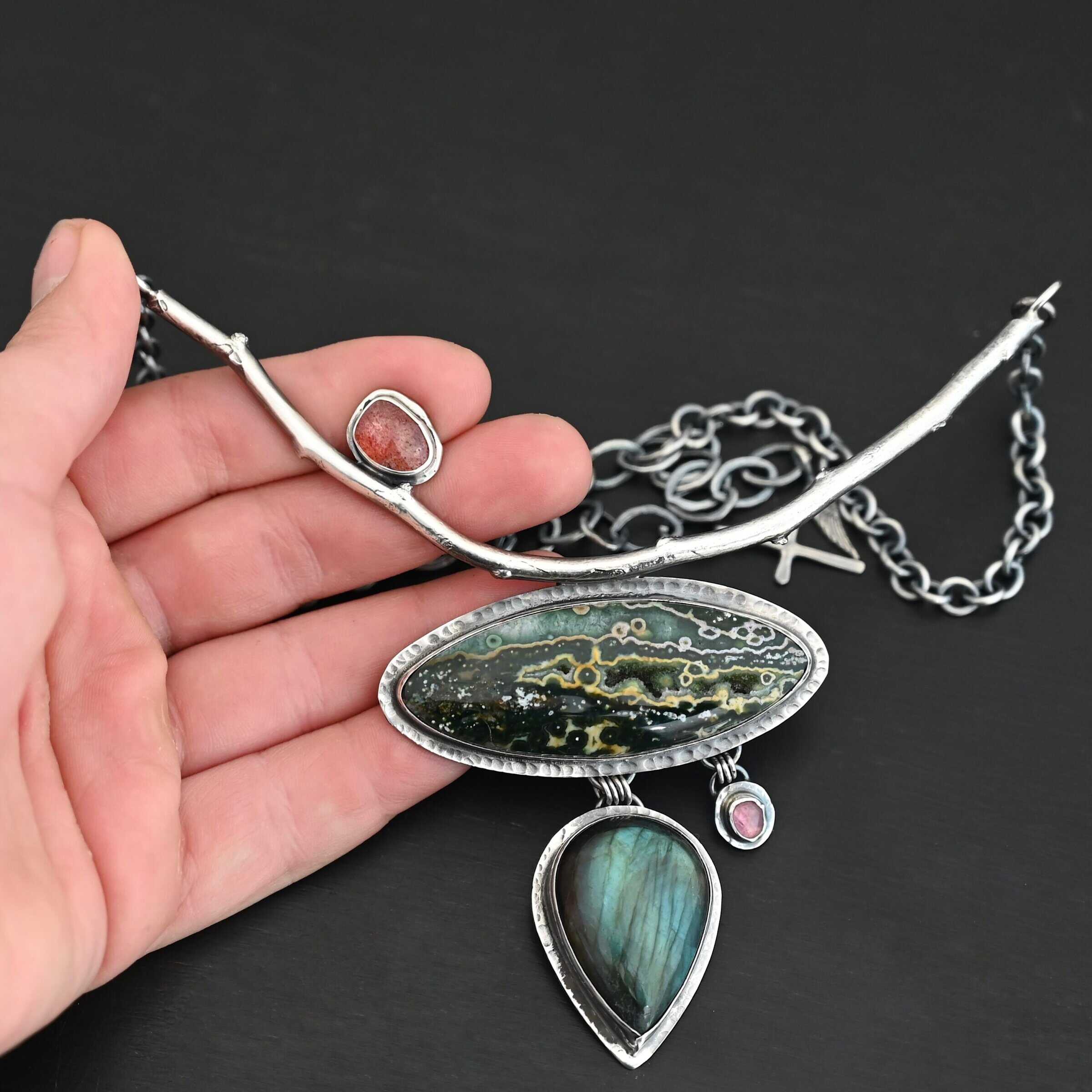 In The Darkness, There Is Light Necklace "In The Darkness, There is Light" Necklace. There is a large, curved, sterling silver casted twig. Soldered beneath it is a horizontal, oval ocean jasper. There is a teardrop labradorite hanging beneath that, as well as a small pink tourmaline next to that. There is another oval stone soldered to the top left side of the branch.