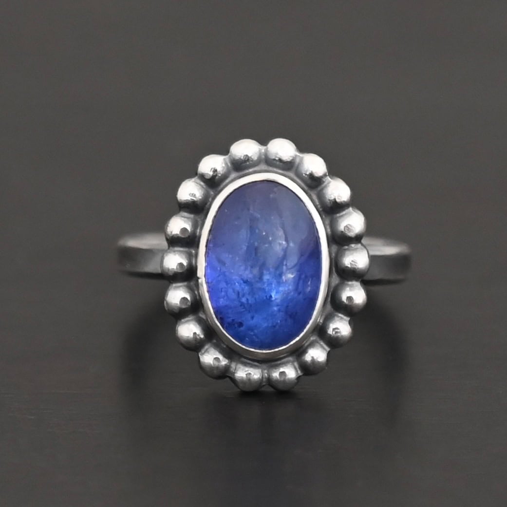 "Asena" Ring. Sterling Silver & Tanzanite. There is an oval, vertical tanzanite in the center with balls of silver circling all the way around it. Ring band is smooth & lightly oxidized.
