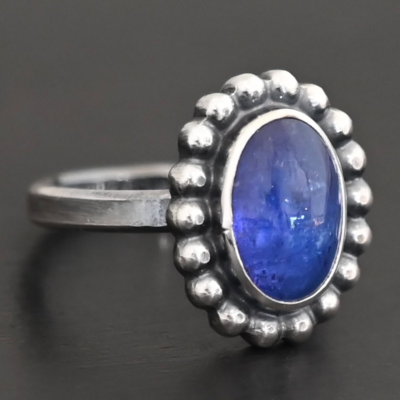 "Asena" Ring. Sterling Silver & Tanzanite. There is an oval, vertical tanzanite in the center with balls of silver circling all the way around it. Ring band is smooth & lightly oxidized.