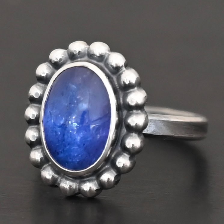 "Asena" Ring. Sterling Silver & Tanzanite. There is an oval, vertical tanzanite in the center with balls of silver circling all the way around it. Ring band is smooth & lightly oxidized.