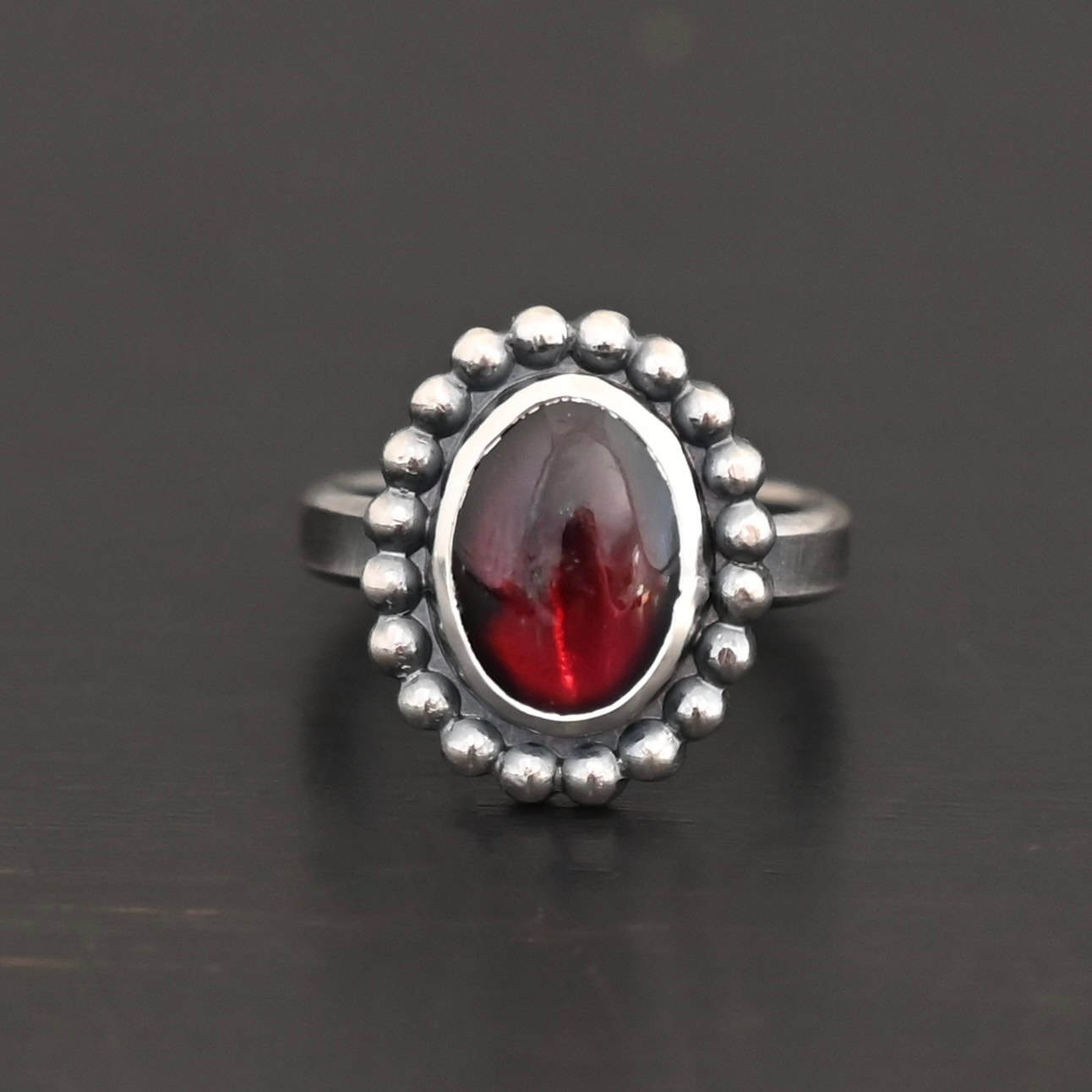 Deep Satin Ring "Deep Satin" Ring. Sterling Silver & Garnet. There is an oval, vertical garnet in the center with balls of silver circling all the way around it. Ring band is smooth & lightly oxidized.