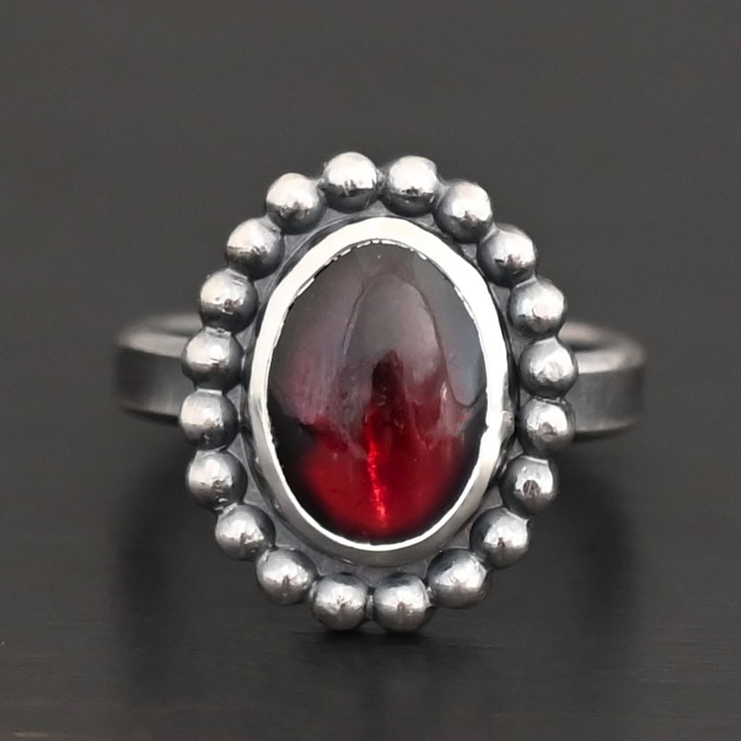 Deep Satin Ring "Deep Satin" Ring. Sterling Silver & Garnet. There is an oval, vertical garnet in the center with balls of silver circling all the way around it. Ring band is smooth & lightly oxidized.