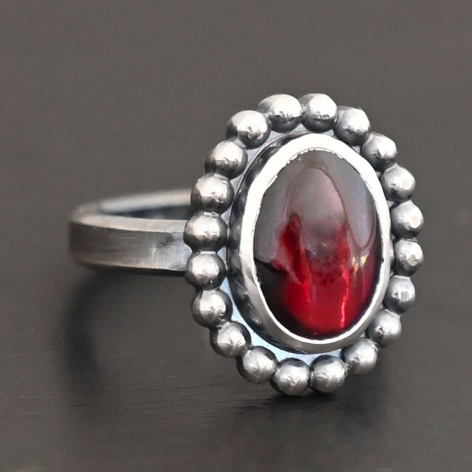 Deep Satin Ring "Deep Satin" Ring. Sterling Silver & Garnet. There is an oval, vertical garnet in the center with balls of silver circling all the way around it. Ring band is smooth & lightly oxidized.