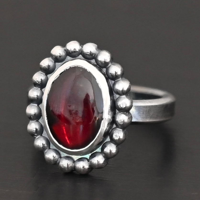Deep Satin Ring "Deep Satin" Ring. Sterling Silver & Garnet. There is an oval, vertical garnet in the center with balls of silver circling all the way around it. Ring band is smooth & lightly oxidized.